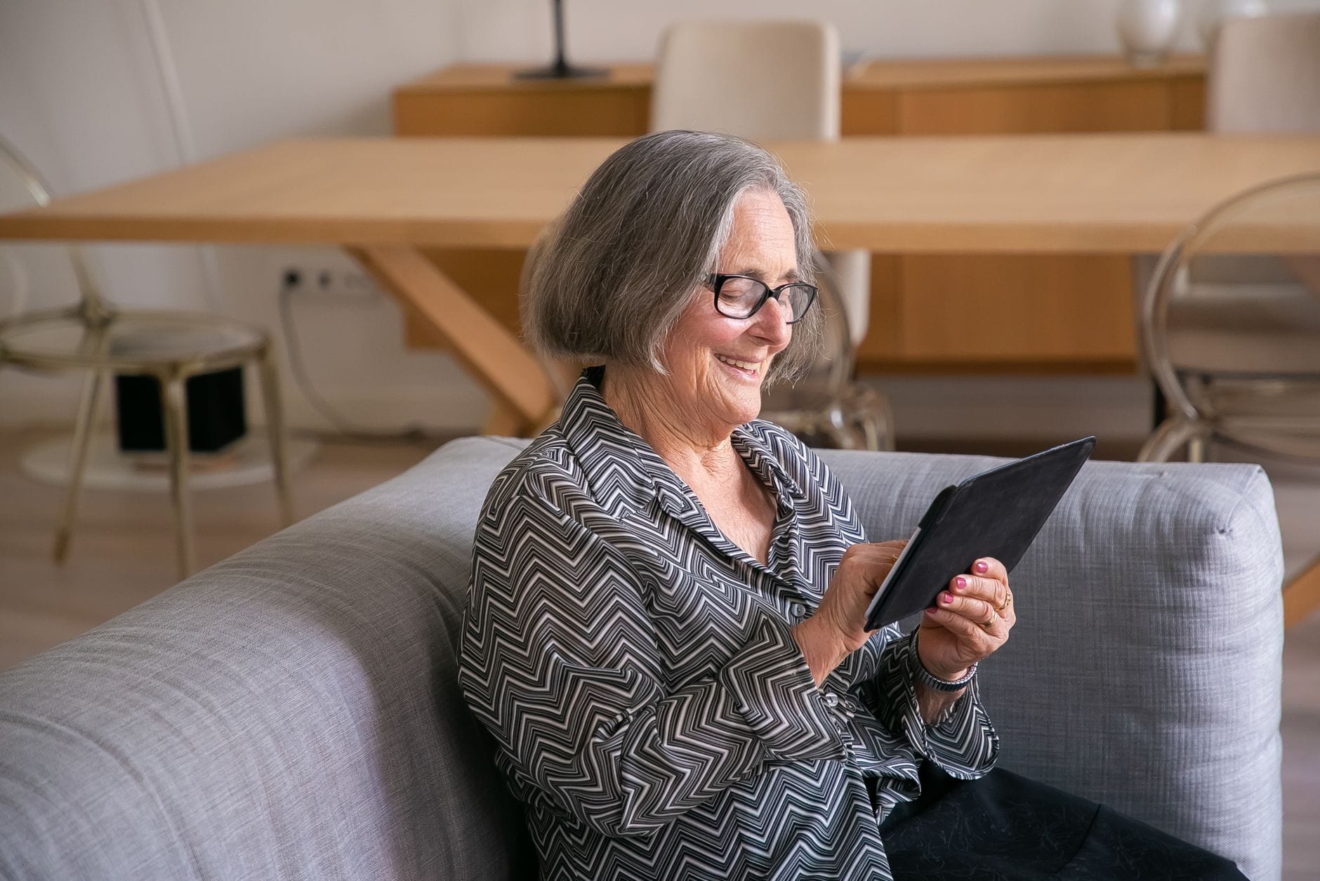 Elderly woman smiling while using a tablet in a cozy living room setting.